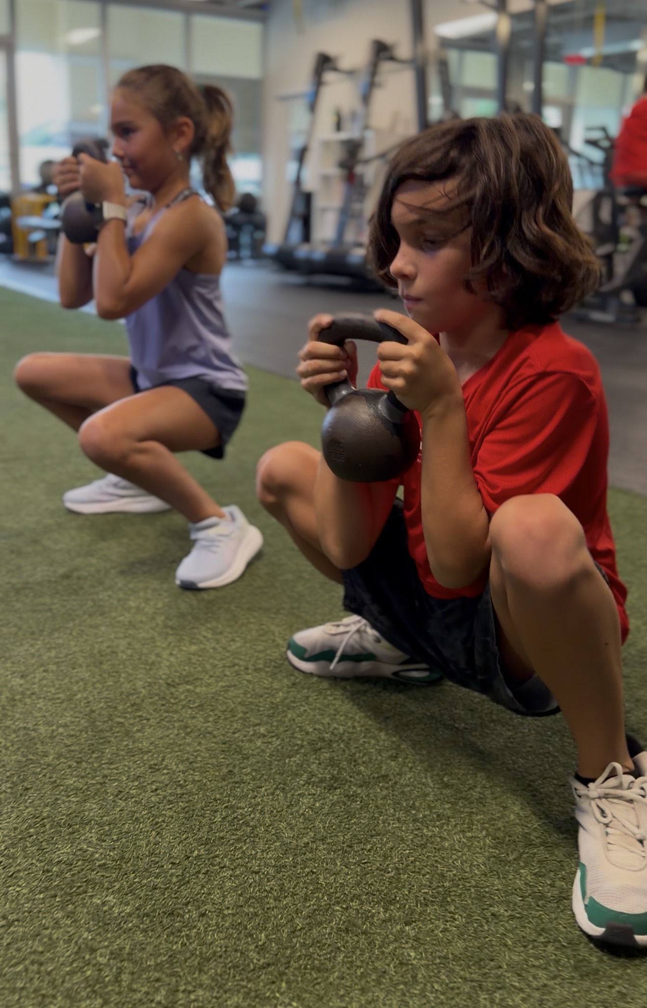 Two kids holding kettlebells in a squat on green turf during Boca Youth Fit