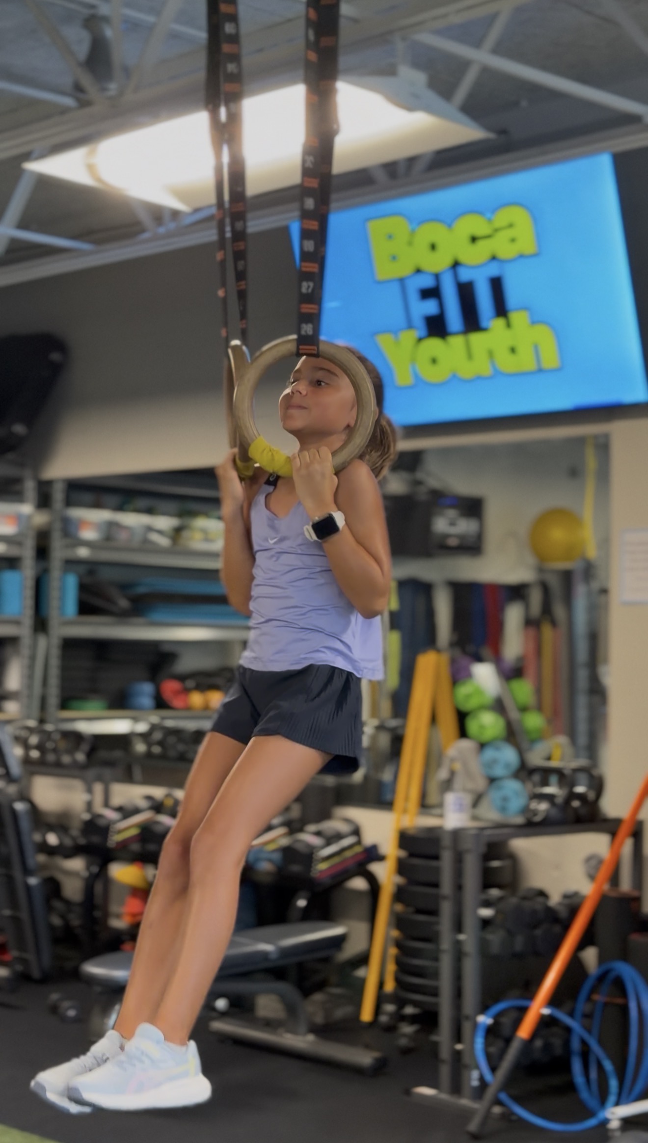 Girl hanging from gymnastic rings in class, Boca Youth Fit on the gym screen behind her